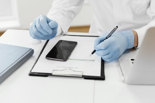 Researcher wearing gloves reviewing clinical trial data on a clipboard beside a smartphone and laptop in a laboratory setting.